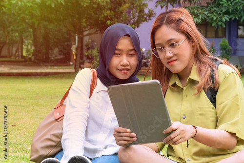Two female college students studying together outdoors on campus, using a digital tablet. Concept of friendship, digital learning, and university life.