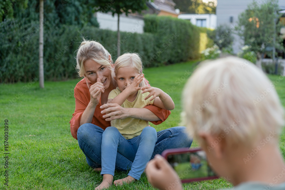 Boy photographing mother and sister gesturing through smart phone in garden Boy photographing mother and sister gesturing through smart phone in garden