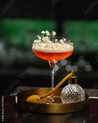 blush-toned cocktail with a frothy top is elegantly garnished with delicate white baby's breath flowers, presented on a golden tray alongside a vintage-style perfume atomizer