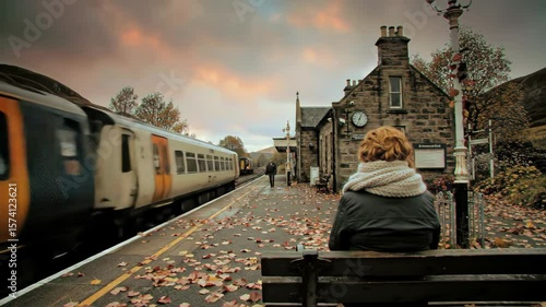 Woman waits on train platform in rural autumn landscape.