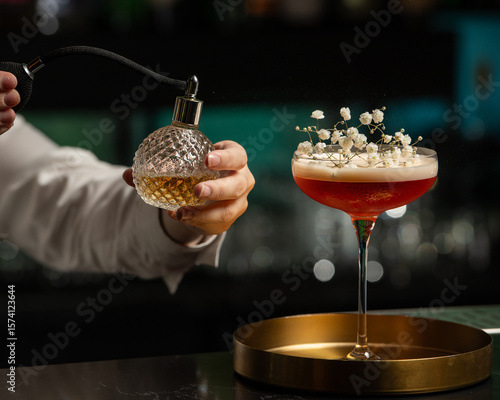 A bartender's hand is delicately misting a blush-toned, floral-garnished cocktail with a vintage perfume atomizer, adding an aromatic layer to the drink's presentation.