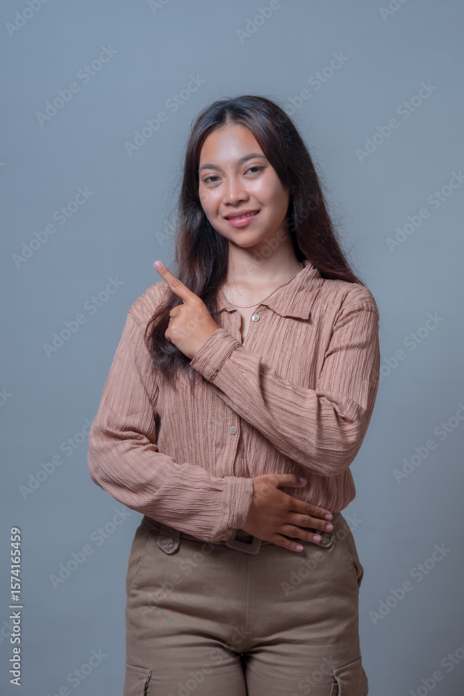 An Indonesian woman in a beige blouse standing confidently with one hand on her waist and the other pointing upwards. Smiling expression, studio shot with a gray background. An Indonesian woman in a beige blouse standing confidently with one hand on her waist and the other pointing upwards. Smiling expression, studio shot with a gray background.