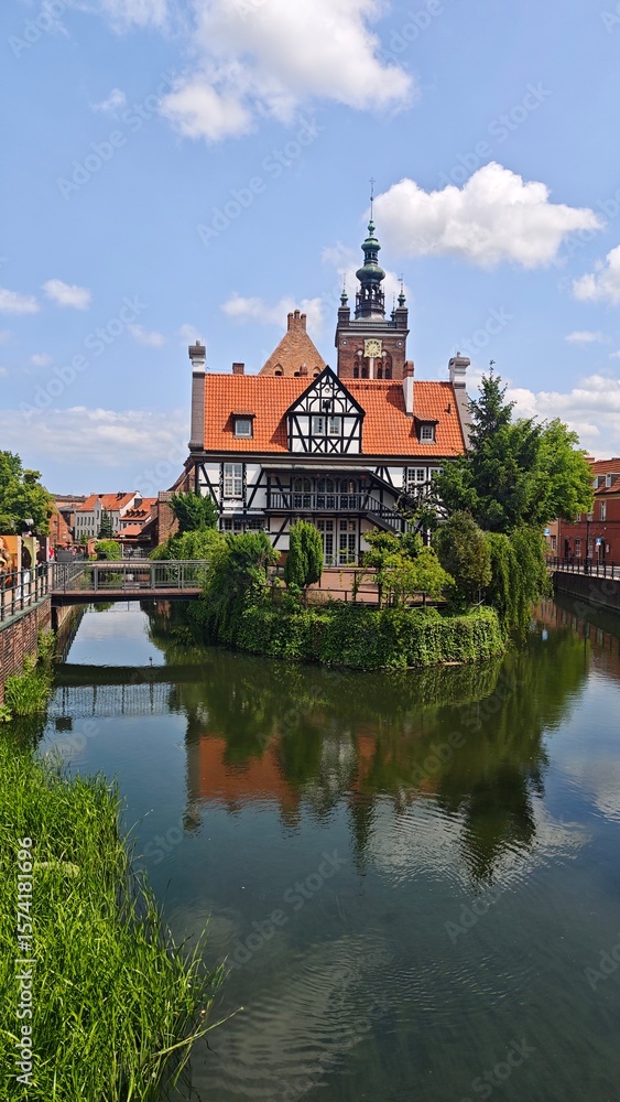 Historic half-timbered building with red tile roof and clock tower reflected in a calm river in Gdansk Historic half-timbered building with red tile roof and clock tower reflected in a calm river in Gdansk