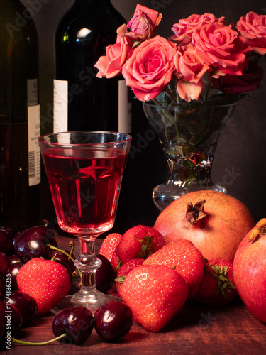 Still life with rose flowers, summer fruits, bottles of wine and a glass of wine. Pomegranate, cherry, sweet cherry, strawberry and watermelon. Alcohol. The still life is stylized as an old painting.