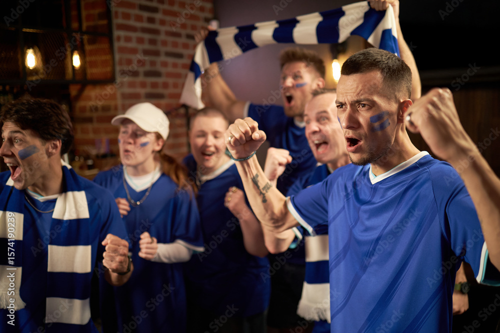 Fototapeta premium Group of diverse young soccer fans cheering and celebrating soccer match victory in championship, wearing blue jerseys and scarves, showing excitement and team spirit