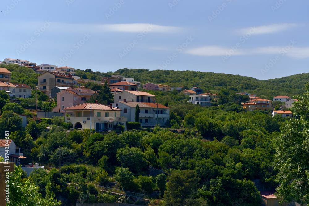 Obraz premium A vibrant cluster of houses with terracotta roofs nestled amidst a dense, green hillside, under a bright blue sky, showcasing a typical Mediterranean residential landscape.