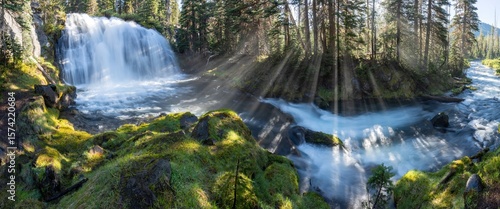 Wild mountain river with a waterfall in the canyon. The sun shines through the trees into the mist and water splinters, creating beautiful rays. A summer morning with a blue sky.
