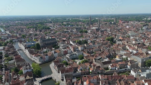 Bruges cityscape from above, showcasing serpentine canals threading between medieval structures, historic rooftops gleaming under bright summer sunlight
