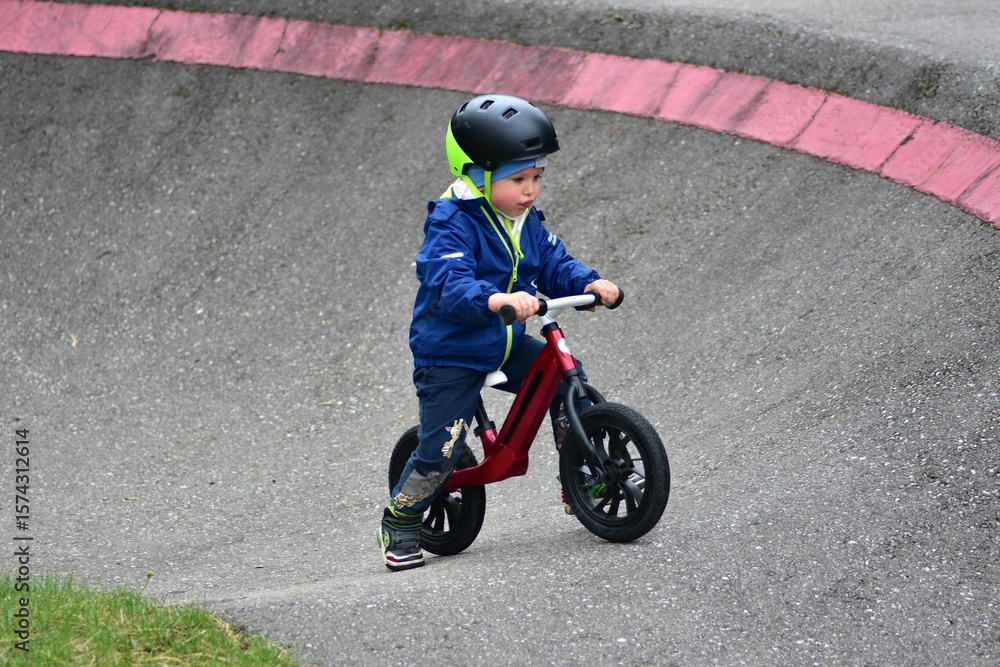 Obraz premium Active young boy in blue jacket and helmet riding a red balance bike on an outdoor pump track. Child practicing cycling skills in green park on summer day. Concept of active childhood and sport.