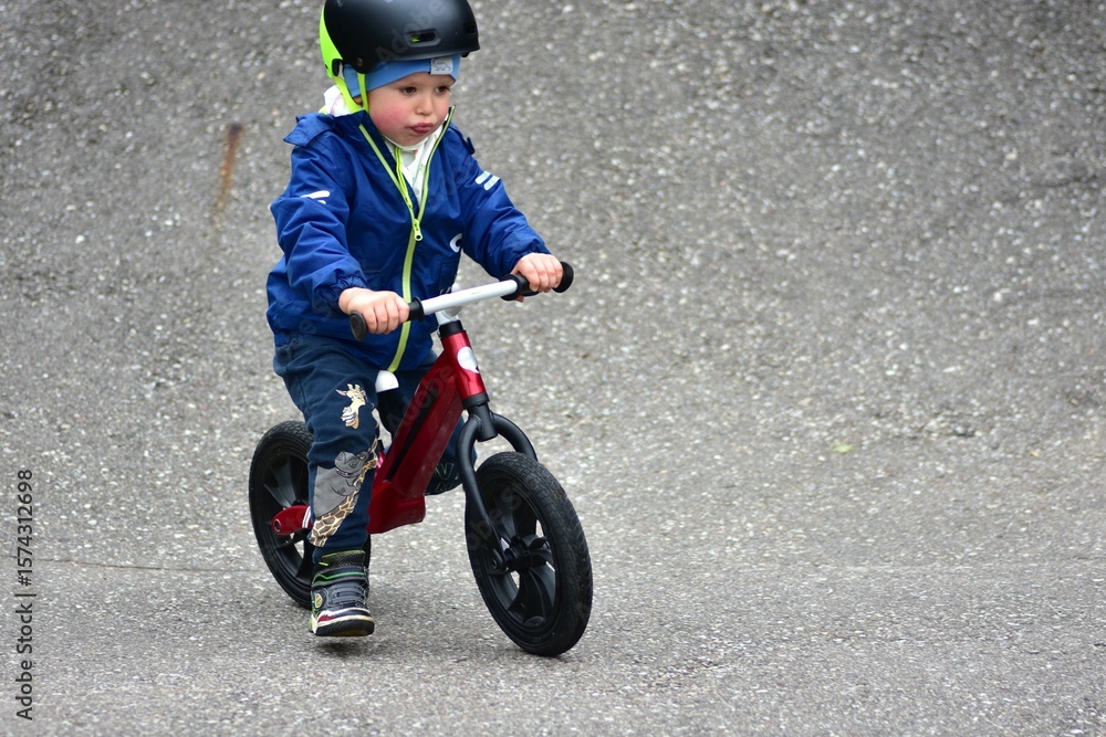 Obraz premium Active young boy in blue jacket and helmet riding a red balance bike on an outdoor pump track. Child practicing cycling skills in green park on summer day. Concept of active childhood and sport.