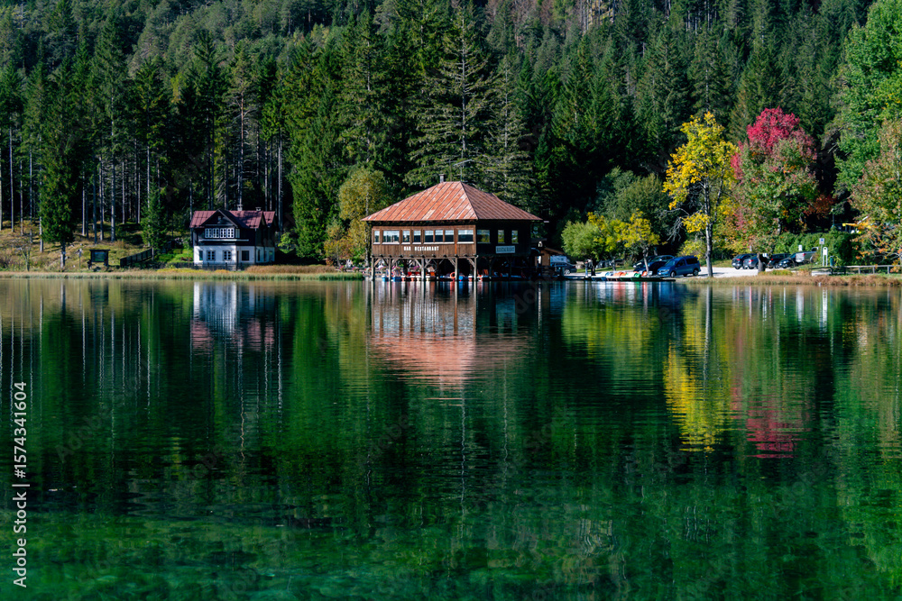 Obraz premium Charming lakeside retreat at Lago di Dobbiaco, Italy. A beautiful wooden building nestled by vibrant forests, creating stunning reflections on the clear green water. Peaceful setting.