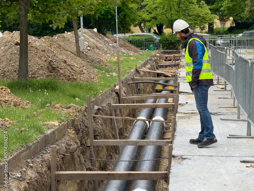 Female engineer in protective gear inspects district heating pipeline installation in urban green zone. Ideal for: energy infrastructure, female engineers, civil works documentation, editorial use.