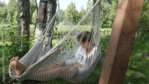 Girl relaxing in hammock. View of cute teenage girl in white t-shirt swinging in hammock at forest camping. Teenager using smartphone.