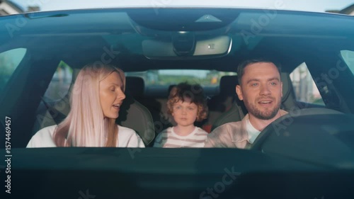 Engaged blonde woman talking to focused man steering calmly through city. Curly-haired boy in back holding teddy bear watching parents. Sitting together during relaxed evening family ride.