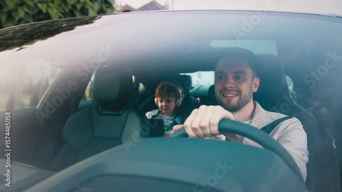 Focused man turning head slightly while steering wheel and cruising through neighborhood. Curious curly-haired boy in car seat wearing headphones and watching video on tablet during relaxed drive.