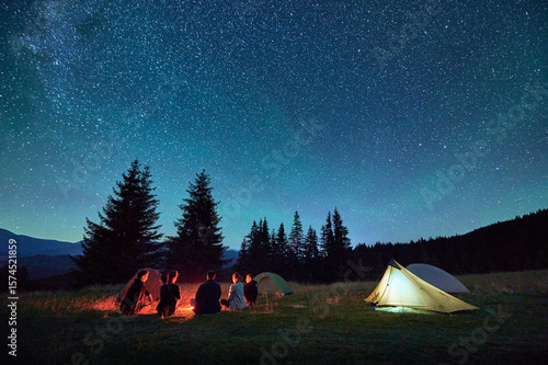 Group of friends hikers resting around campfire, surrounded by illuminated tents under stunning starry sky. Milky Way stretches above, creating magical atmosphere in peaceful mountain wilderness.