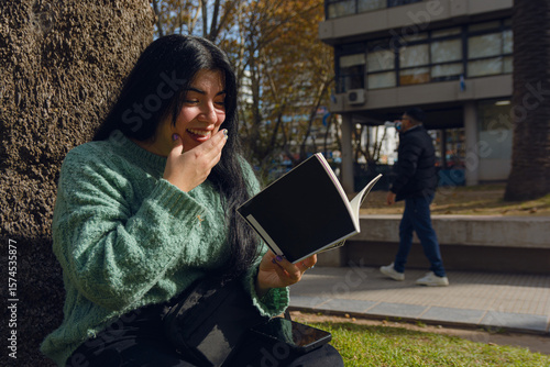 young woman reading a book smiling happily with one hand on her mouth