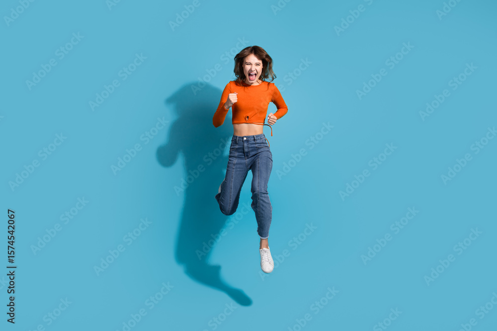 Obraz premium Joyful young woman in stylish orange top and jeans leaping passionately in front of a vibrant blue background full of energy and charm.