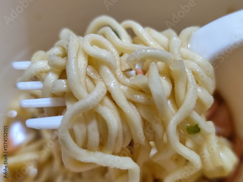 
Gulungan mi instan putih yang kenyal diangkat dengan sendok dari mangkuk, menonjolkan tekstur dan kelezatannya, dengan latar belakang mangkuk yang buram.
