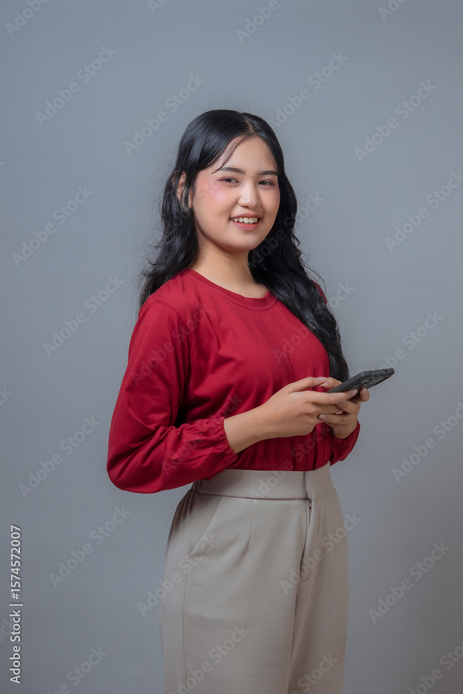 Cheerful Indonesian woman in red blouse and beige pants holding a smartphone while smiling confidently. The modern look and tech gesture reflect connection, lifestyle, and youth. Cheerful Indonesian woman in red blouse and beige pants holding a smartphone while smiling confidently. The modern look and tech gesture reflect connection, lifestyle, and youth.