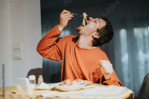 Obraz na płótnie A young man appears to relish a delicious pasta meal while sitting at a table in the cozy ambiance of a home kitchen