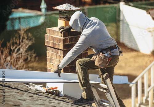 Canvas Print A roofer in a hoodie is working on a roof, installing or repairing a section near a chimney with various tools