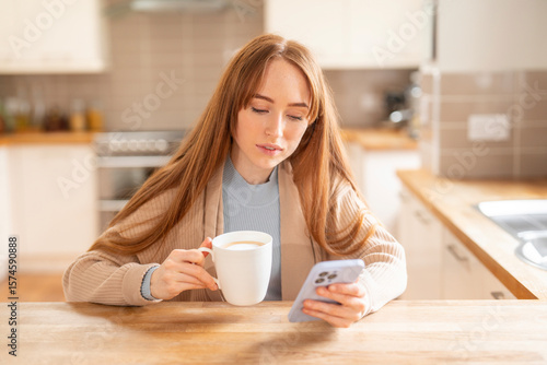 Принти на полотні Woman enjoys a warm drink while browsing on her smartphone in a cozy kitchen dur