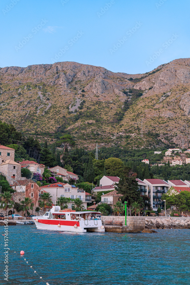 Obraz premium Red and white catamaran sailing near traditional coastal town with mountains rising behind. Summer tourism image.