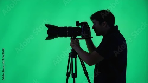 Silhouette of person holding DSLR camera, adjusting lens and snapping photos in different directions, standing before a chroma green background, creative industry vibe