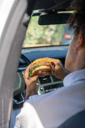 Fototapete Zoom sur un homme assis dans sa voiture tenant un burger dans sa main, prêt à manger, dans une ambiance urbaine et décontractée