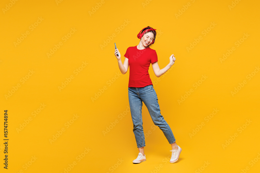 Fototapeta premium Full body young woman she wearing red t-shirt casual clothes listen to music in headphones holding in hand use mobile cell phone isolated on plain yellow background studio portrait. Lifestyle concept.