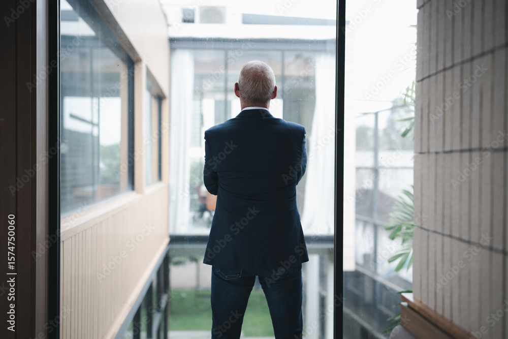 Senior executive is looking outside the window of his contemporary office building with his arms crossed, contemplating the view and thinking about future business decisions Senior executive is looking outside the window of his contemporary office building with his arms crossed, contemplating the view and thinking about future business decisions