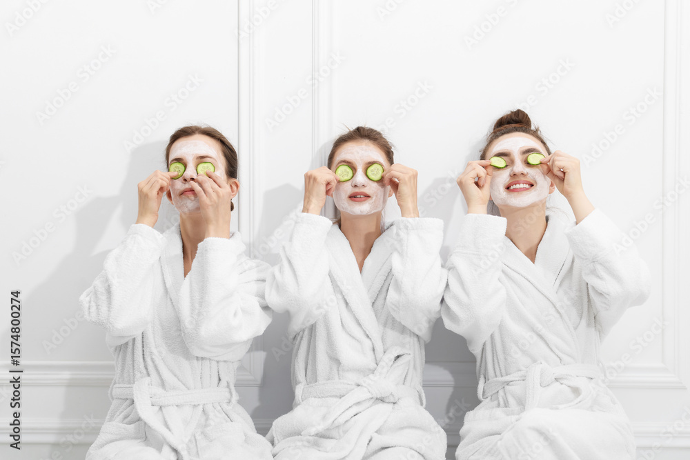 Obraz premium Happy women with facial masks and cucumber slices in bathrobes after spa procedures near white wall