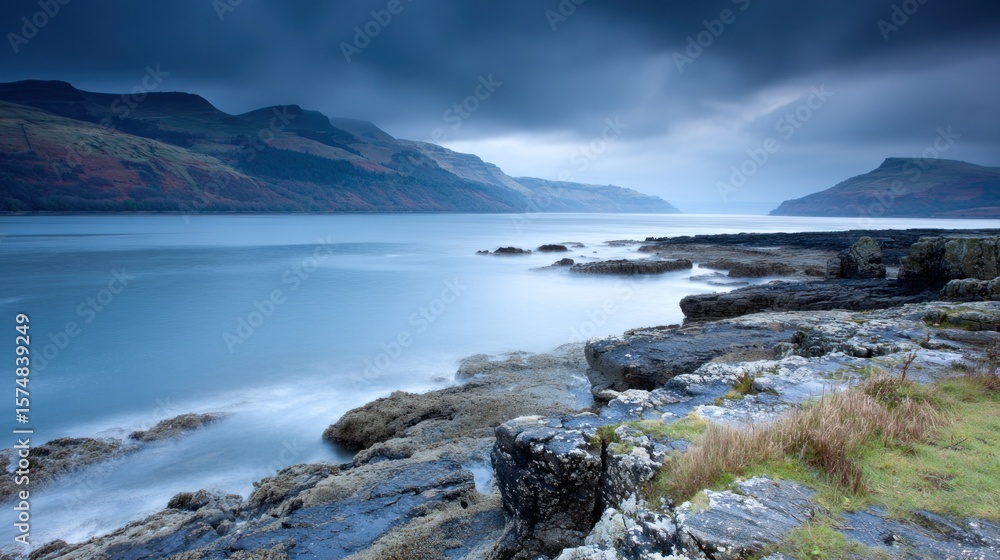 Fototapeta premium Serene coastal scene with calm waters, dark rocky shoreline, and distant mountains under a moody sky