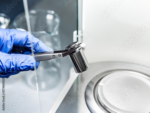 A operator's hand is holding steel calibration weight to place on the analytical balance.