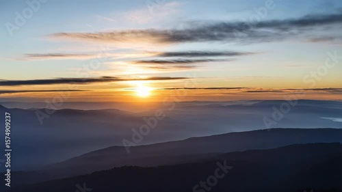 A tranquil sunrise over misty mountains, with golden light illuminating the horizon and soft clouds scattered in the sky.