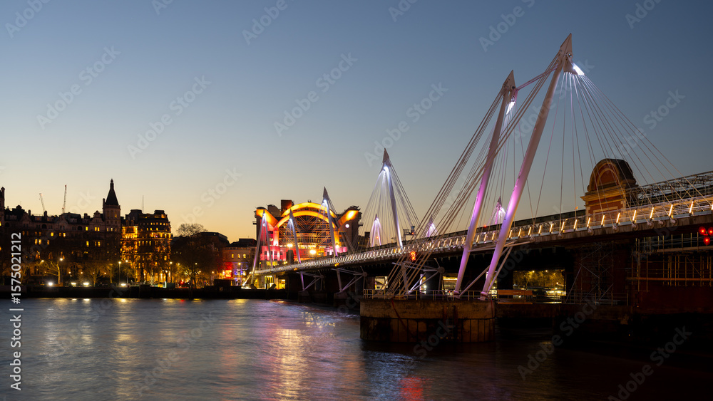Obraz premium The illuminated Charing Cross Bridge spans the River Thames in London during twilight. Surrounding lights reflect on the water, creating a peaceful evening atmosphere for viewers.