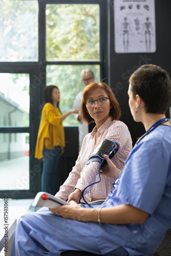 Asian woman does health check with nurse monitoring blood pressure with electronic device, checking vitals and doing a routine evaluation in modern clinic lobby. BP monitor control.