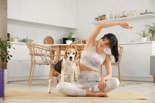 Young sporty woman with cute Beagle dog practicing yoga on mat in kitchen