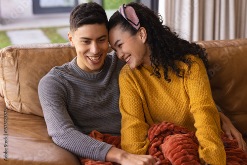 Couple snuggling under red throw blanket on brown leather couch in living room by large window