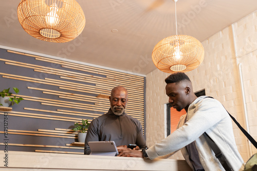 African American staff member and visitor examining tablet at counter under rattan lamps in lobby