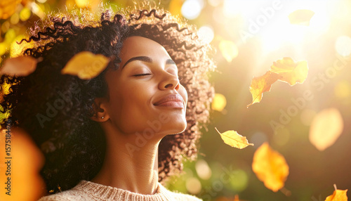 A woman with curly hair enjoys the autumn season, eyes closed, as leaves fall around her in the sunlight.