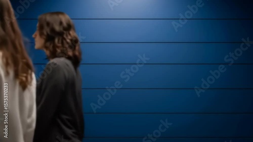 Two women walking past a dark blue wall in a modern setting, ideal for fashion and lifestyle promotions