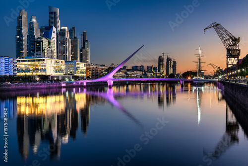 Aerial view of the lit Puente de la Mujer bridge reflecting in the dark water, with a backdrop of modern skyscrapers, Buenos Aires, Argentina.