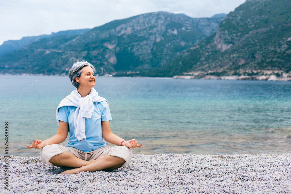 Fototapeta premium Happy mature woman meditating by the sea, enjoying peaceful moment on rocky beach outdoors