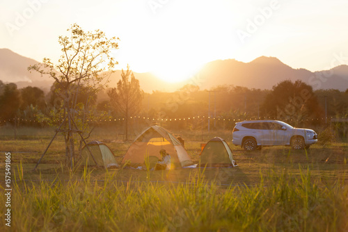A heartwarming camping scene during golden hour with tents, a car, and silhouettes of people enjoying nature under a warm sunset in the open field.