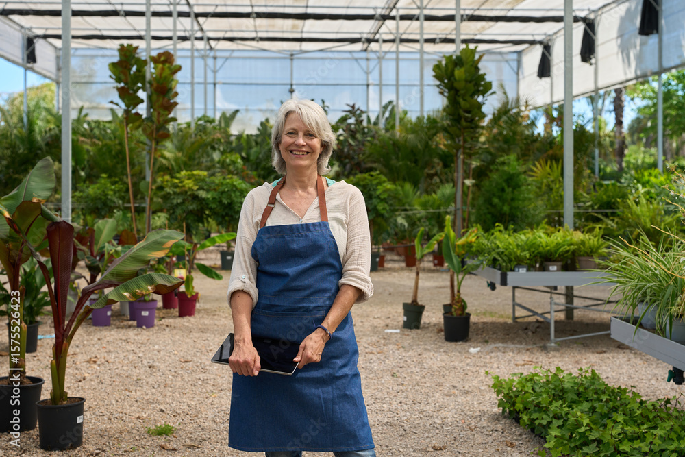 Fototapeta premium Senior woman gardener smiling and holding tablet in greenhouse