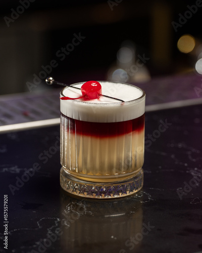 A striking layered cocktail features a rich red top over a pale yellow base, crowned with a thick white foam and garnished with a single vibrant red cherry on a skewer