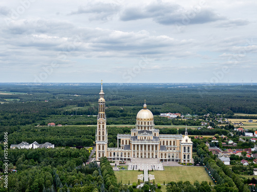 Aerial view of the Basilica of Our Lady of Lichen surrounded by lush forest, lake, and rural landscape under clear blue summer sky. Shot in Poland