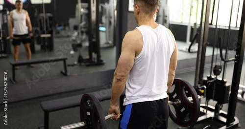 Strong man in activewear lifts barbell with shoulders in gym backside view. Skilled bodybuilder trains muscles working with large weight near mirror